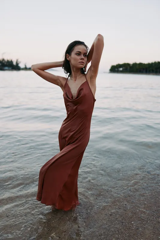 Stylish woman stands in shallow sea water wearing a rust red dress, posing with arms raised, soft natural light, calm shoreline, graceful posture, fashion portrait with minimal background and gentle
