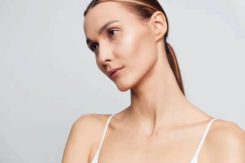 A close-up portrait of a young woman with flawless skin and minimal makeup looks thoughtfully to the side against a neutral grey background. Concept for beauty and skincare.