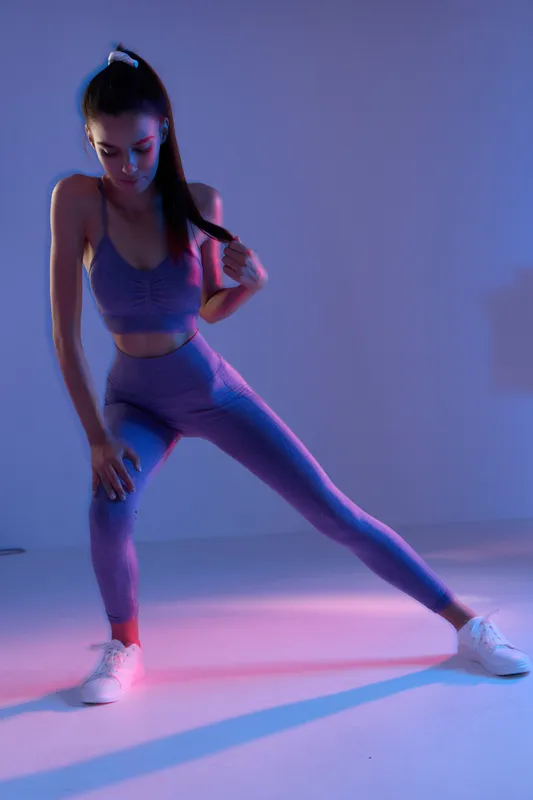 Young woman sport fitness stretching in purple activewear with dynamic lighting indoors, focused on warm-up and flexibility in a modern studio setting.