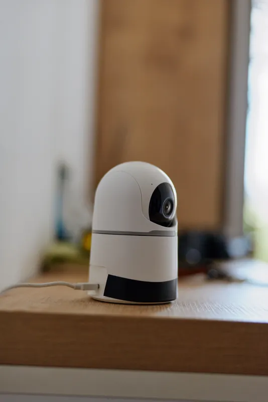 Compact indoor security camera sits on a wooden table in a softly lit room, presenting a minimalist home surveillance concept with a modern, unobtrusive presence