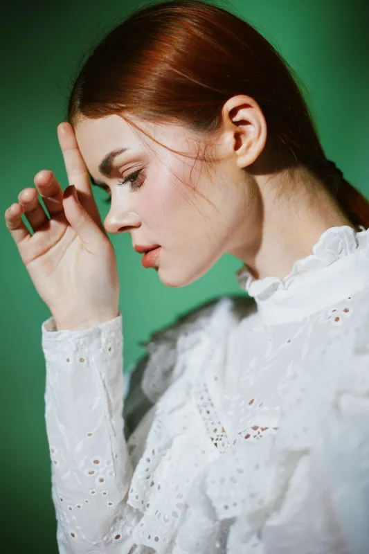 Portrait of a young woman in a white lace dress with a thoughtful expression against a green background, showcasing elegance, emotion, and seasonal style
