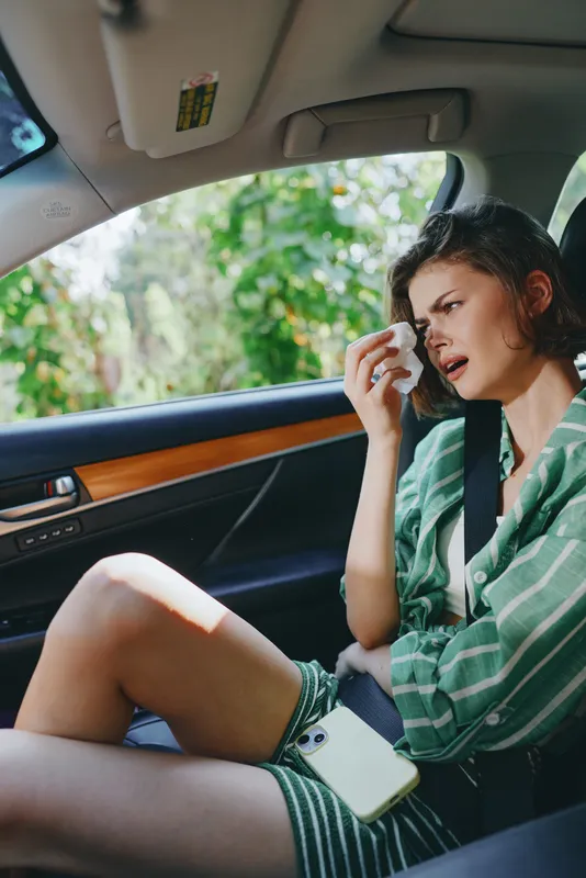 A young woman sits in a car, wearing a striped green shirt and seatbelt, with a pensive expression, capturing a quiet, introspective moment inside the vehicle.