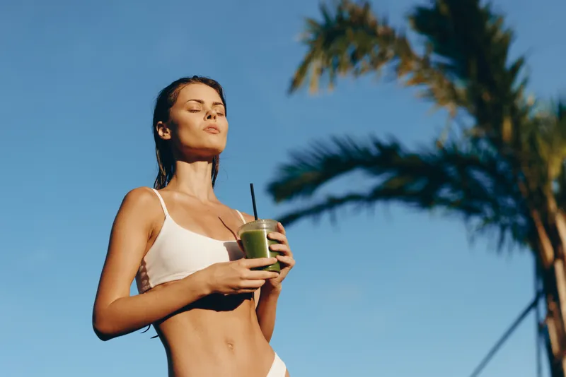 Bright outdoor scene featuring a woman in a bikini holding a green smoothie, sunlit skin, eyes closed, relaxed pose under a clear blue sky with palm trees, conveying healthy lifestyle and summer vibe