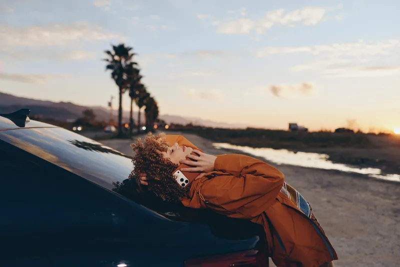 Woman in lifestyle portrait, curly hair leaning on car hood at sunset near ocean shoreline, wearing orange jacket, hands touching hair, warm golden light, scenic road background, outdoors
