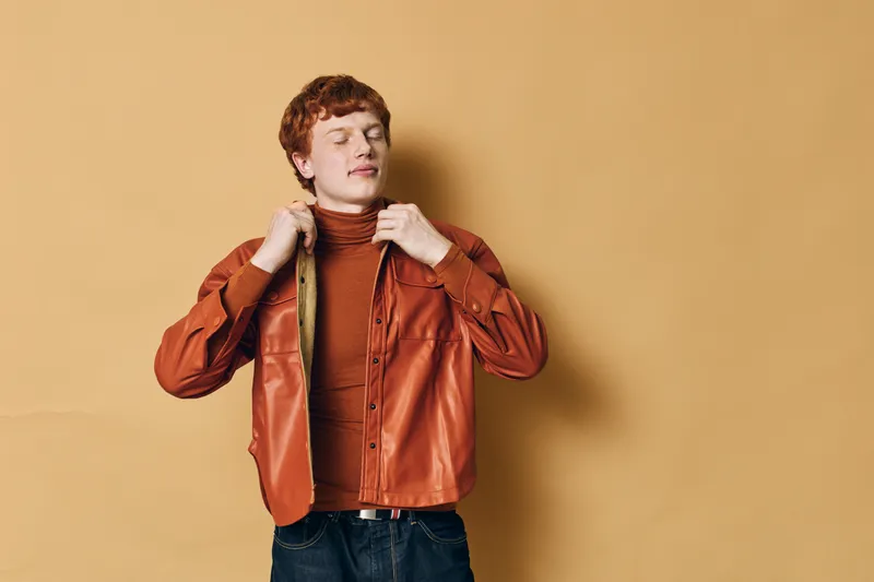 Young man with curly red hair adjusting brown leather jacket over turtleneck sweater, standing against beige background with closed eyes, expressing calm and confidence.
