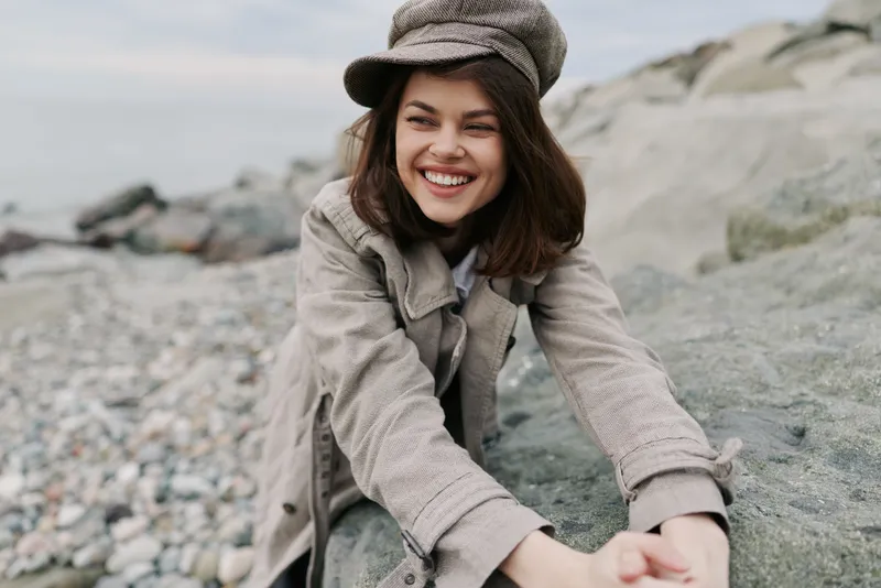 Smiling young woman in beige coat and hat leans on rocks at pebble beach. Natural outdoor scene with casual fashion, autumn mood, and positive lifestyle concept.