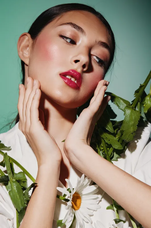 Close-up of a woman with glossy red lips and flawless skin posing with green foliage and white flowers on a pastel green backdrop. Beauty and fashion creative portrait concept.