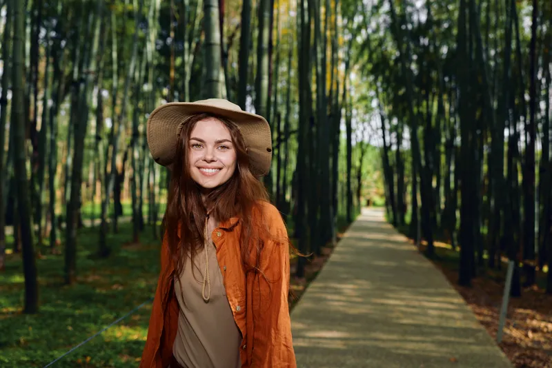 Woman smiling in a straw hat walking in an autumn park on a tree lined path, portrait of a happy young adult wearing a cozy jacket with natural light and seasonal foliage.
