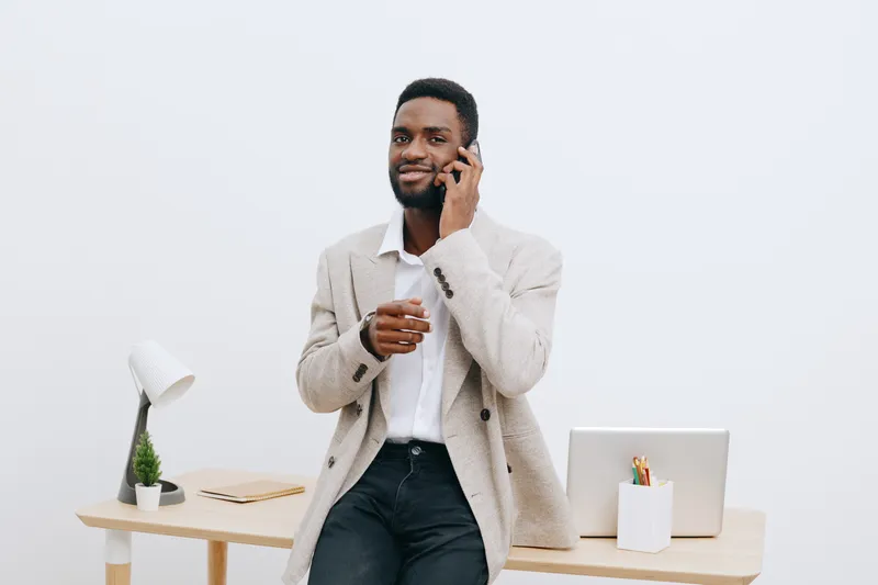 Smiling man working in modern office, talking on mobile phone, dressed casually, using laptop and stationery, professional workspace, light minimalistic environment, lifestyle concept, business commun