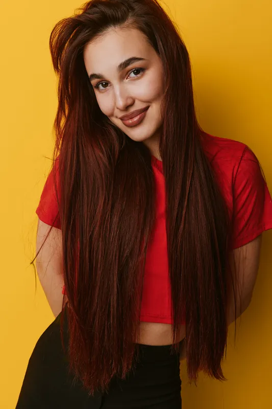 Young woman with long straight hair wearing red cropped t shirt posing smiling against yellow background. Casual style, beauty portrait, confident female with natural makeup and relaxed posture.
