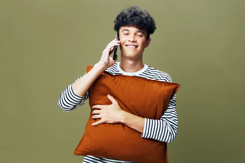 young man talking on phone while hugging rust-colored pillow, smiling with closed eyes, wearing striped shirt, olive green background, cozy casual moment