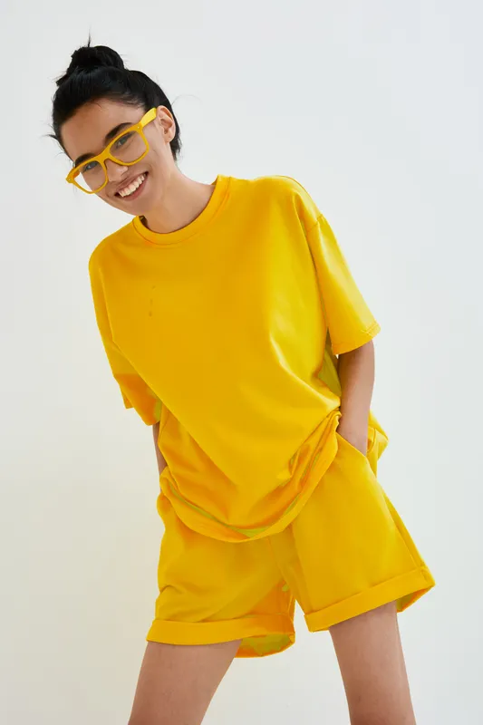 Smiling woman wearing bright yellow casual clothes and matching glasses, posing confidently with hands in pockets against plain white background.