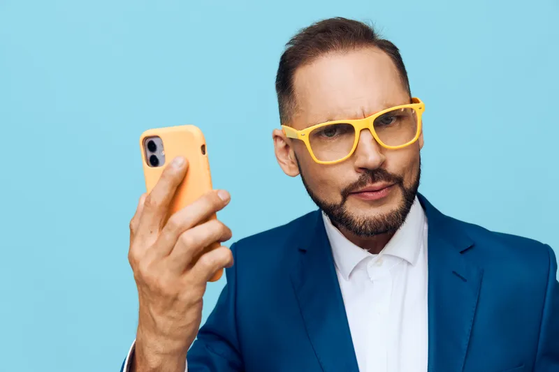 A professional man stands against a bright blue background, wearing a blue suit and yellow glasses, looking at a smartphone with a confident, focused expression.