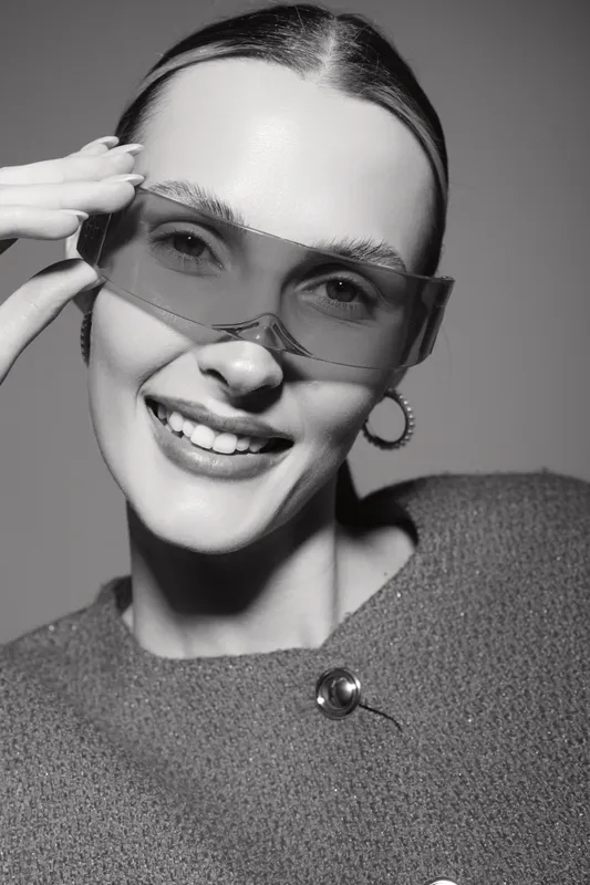 Smiling caucasian woman with fair skin wearing transparent sunglasses and hoop earrings, posing confidently in studio lighting. Concept of fashion, beauty, and style.