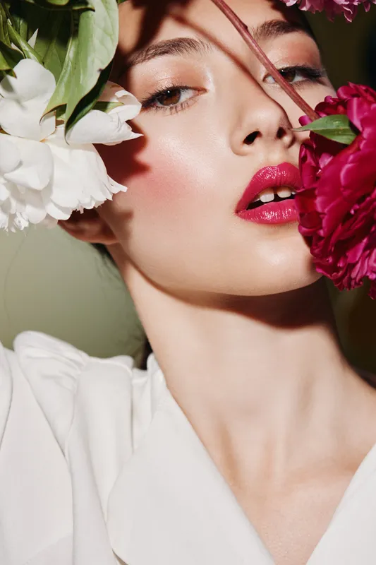 Close-up of a beautiful woman face with radiant skin and glossy pink lips surrounded by vibrant flowers. Creative beauty concept with natural lighting on neutral background.