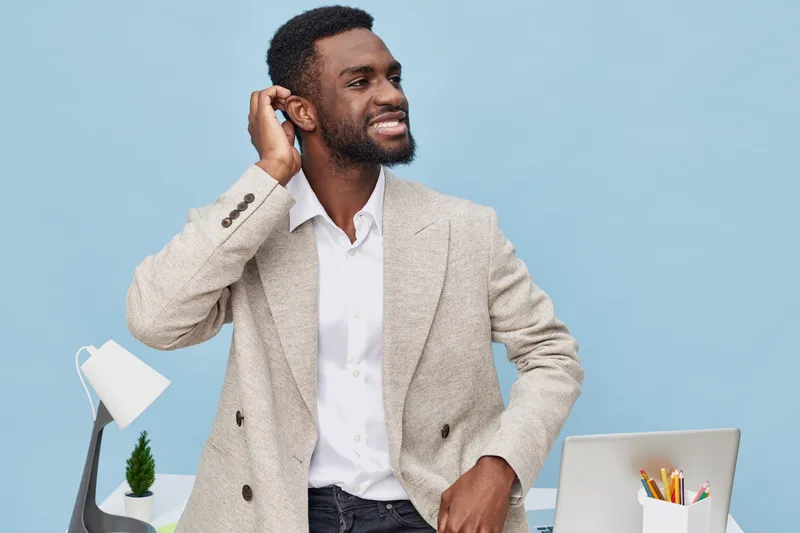 Smiling man with dark skin dressed in beige blazer and white shirt seated at modern workspace with desk lamp and pen holder, looking away happily on solid light blue background, studio portrait. Peopl