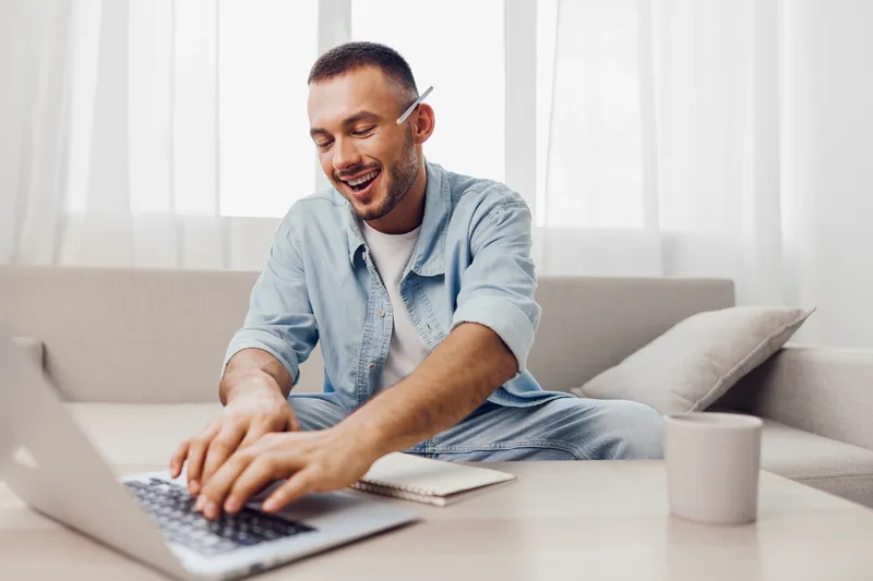 Smiling man working on a laptop at home, wearing casual clothes, seated comfortably with a notebook and a cup, creating a cozy atmosphere for productivity.
