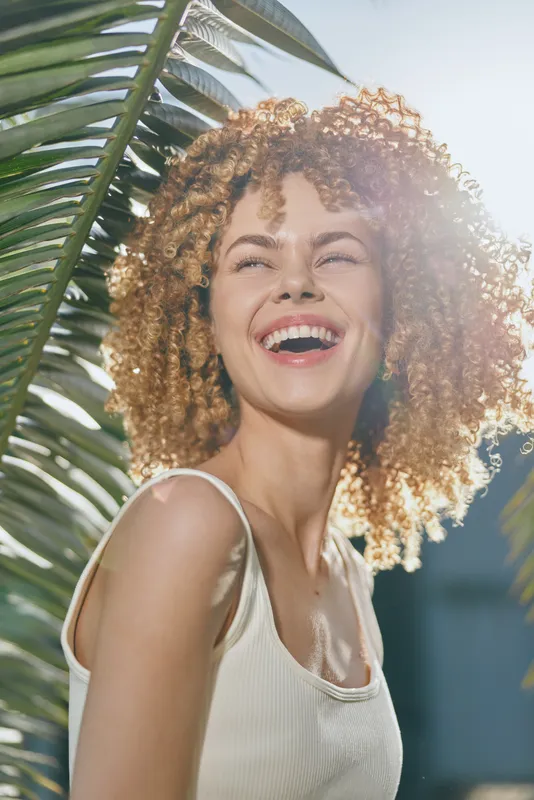 smiling woman with curly hair enjoys sunny day outdoors near palm leaves, wearing white tank top, radiating happiness and natural beauty in bright sunlight