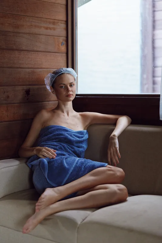 Relaxed woman wrapped in blue towel sitting on beige sofa near window in wooden room, wearing shower cap, natural light highlighting calm expression and cozy atmosphere.