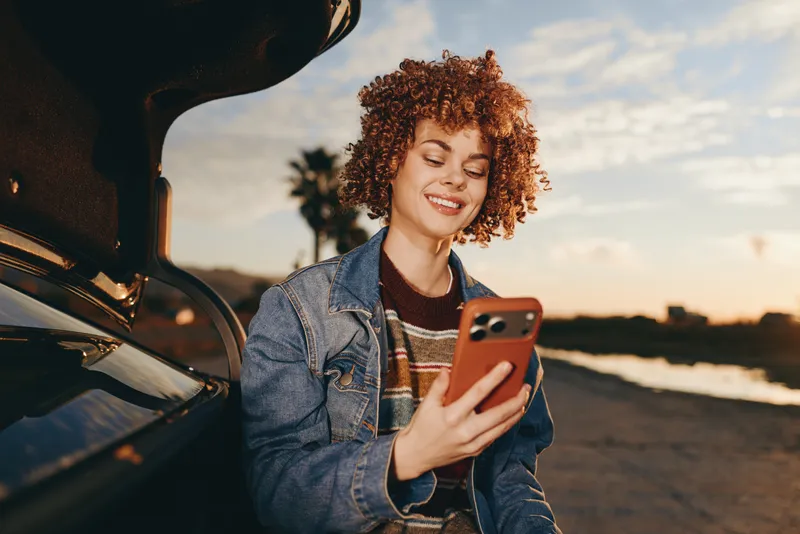 Smiling woman with curly hair holding a smartphone near a car at sunset on a road, Lifestyle travel moment outdoors, warm golden light, casual denim jacket