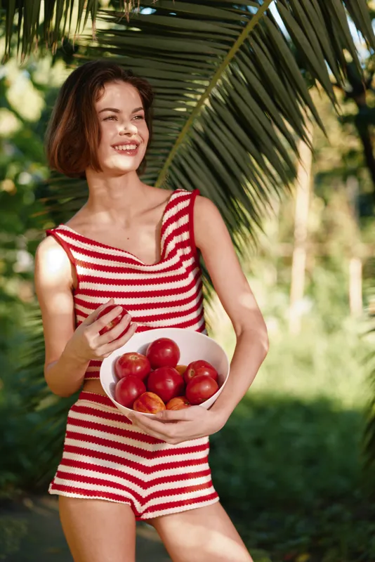 A cheerful woman in a red striped swimsuit holds a bowl of apples outdoors, smiling in a sunny garden. Palm leaves frame the scene, creating a bright, playful summer moment.