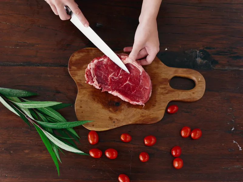 Женщина с ножом в руке режет свежее сырое мясо для жарки на кухне