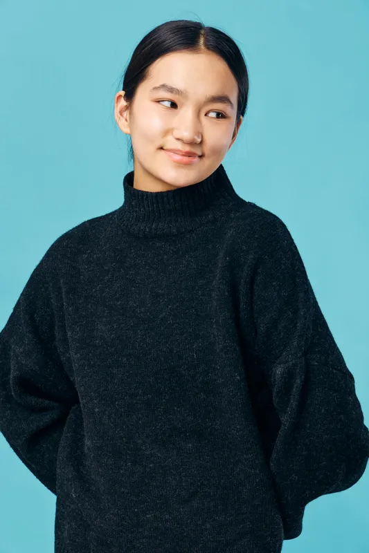 Young woman wearing black sweater posing with hands behind back looking sideways smiling on blue background. Casual style, confident gesture, indoor portrait with relaxed mood and natural light.