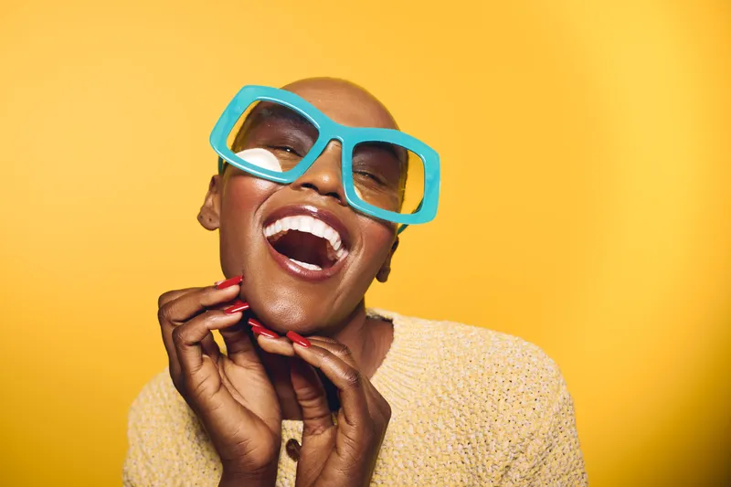Happy black woman with dark skin and blue oversized glasses smiles joyfully on yellow background. Close-up portrait captures positive mood and vibrant fashion style of confident female.