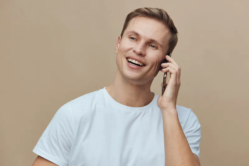 Smiling happy young caucasian man talking on mobile phone wearing white t-shirt light brown background concept digital nomad communication