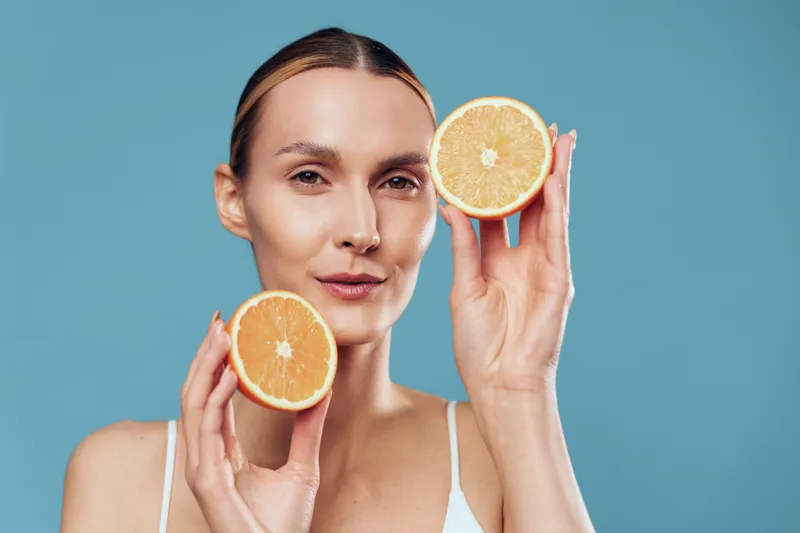 Studio portrait of a female one person with blonde hair holding two orange halves, creating a bright citrus moment against a blue backdrop with a clean minimalist vibe