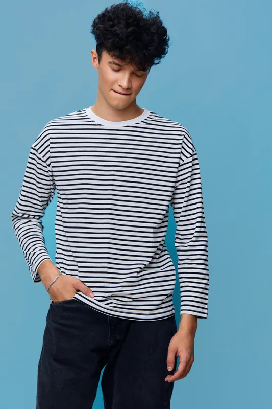 young man in striped shirt stands thoughtfully against blue background with casual style and dark pants, expressing calm and introspective mood in simple portrait
