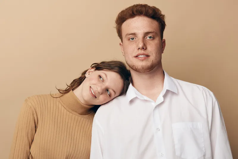 Young couple with woman leaning on man's shoulder, both looking at camera. Casual clothing and neutral background create a calm and intimate portrait of togetherness and affection.