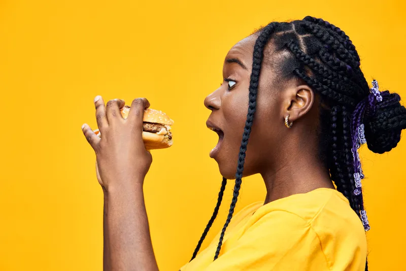 Young Black woman excitedly holding a juicy hamburger against a vibrant yellow background, showcasing her love for fast food in a lively, playful atmosphere