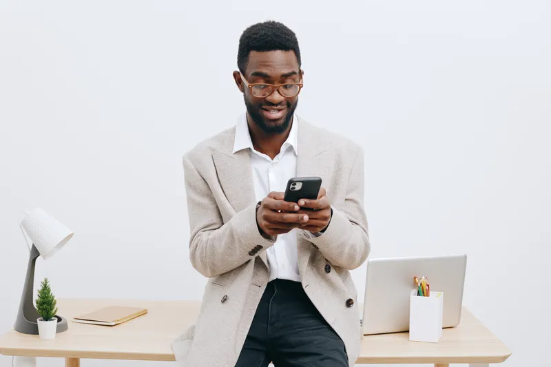 Smiling man with glasses using smartphone in modern office setting, standing near desk with laptop, notebook, and plant, wearing casual beige cardigan and white shirt. People lifestyle concept