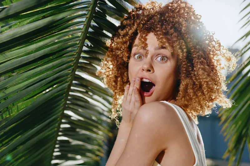 surprised woman with curly hair posing near tropical palm leaves outdoors, bright sunlight highlights her expressive face and natural skin tone