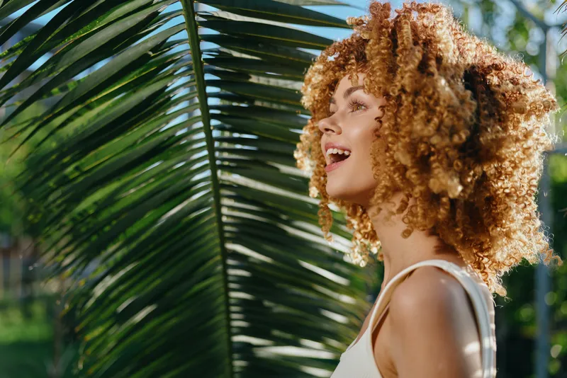smiling woman with curly hair outdoors near palm leaf in sunlight, enjoying nature, summer vibes, natural beauty and happiness expression