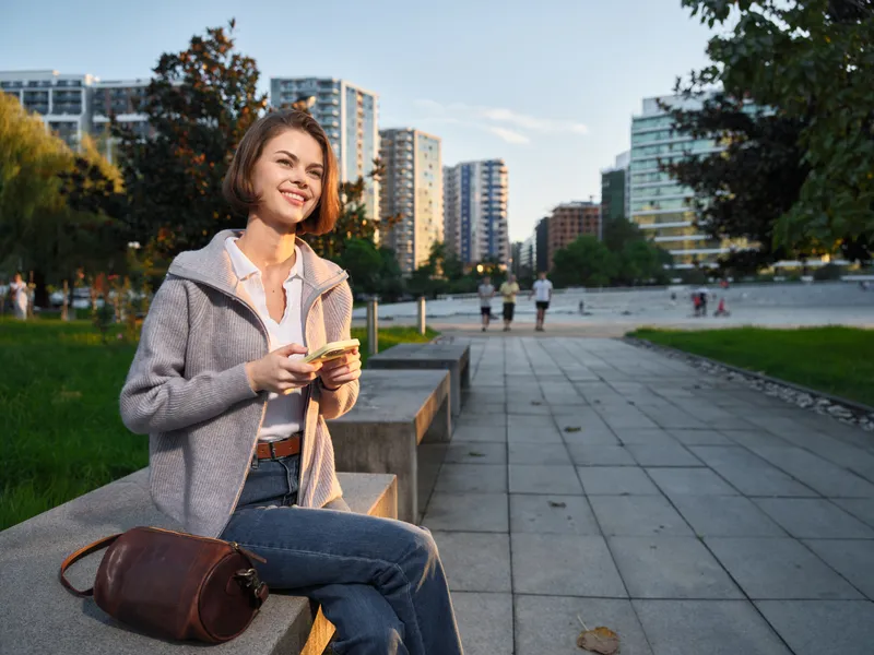 A cheerful woman sits on a park bench in a modern city setting, smiling while looking at her phone. She wears a casual jacket and a leather bag rests nearby, creating a relaxed outdoor atmosphere.