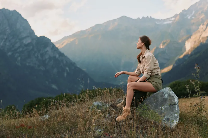 Woman sits on a rock in a mountain landscape, conveying outdoor lifestyle and contemplation, with rugged scenery and a calm, confident presence. Soft lighting enhances warmth