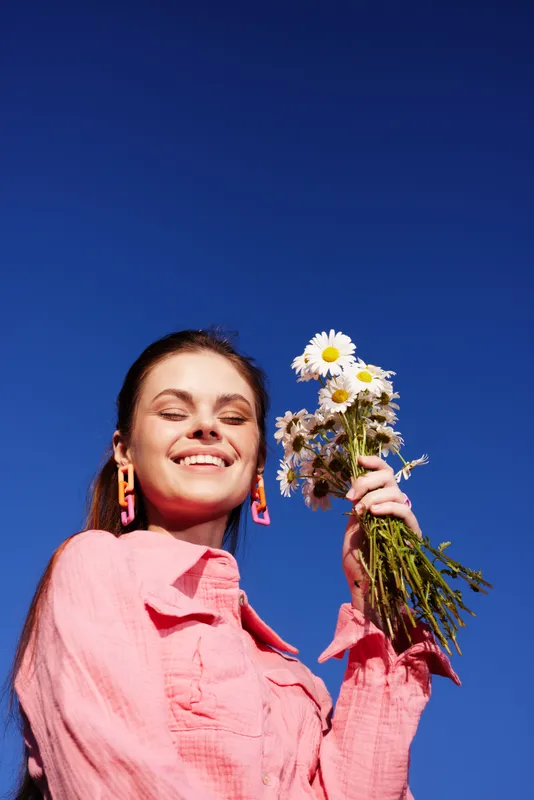 Young woman smiling joyfully while holding wildflowers against a bright blue sky, embodying a carefree summer spirit and natural beauty