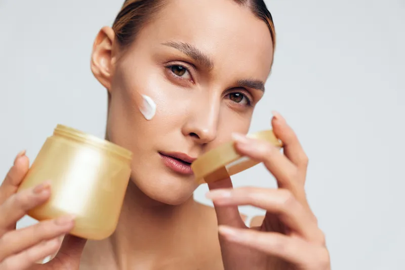 Woman skincare close up, face cream for moisturizing beauty treatment, holding jar and applying with hands, studio gray background, calm expression, soft light
