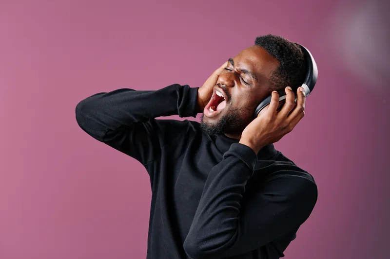 A joyful Black man wearing headphones passionately sings along to music against a purple background, expressing pure happiness and energy
