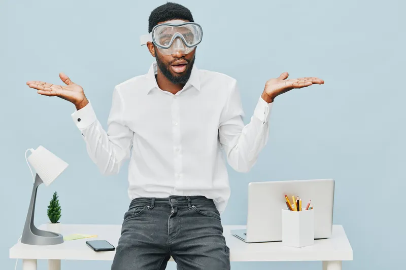 Modern young man with glasses and beard wearing white shirt poses with a confused expression, shrugging his shoulders, in a bright office with minimal decor, laptop, desk accessories, concept of uncer