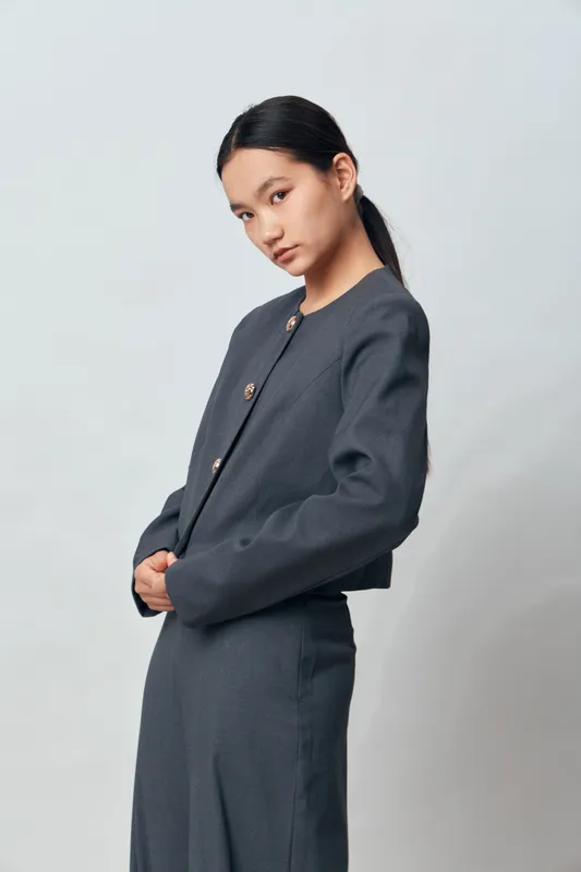 Woman in modern gray suit standing with hands clasped, looking sideways on light background. Stylish professional fashion model with natural makeup and sleek black hair in ponytail.