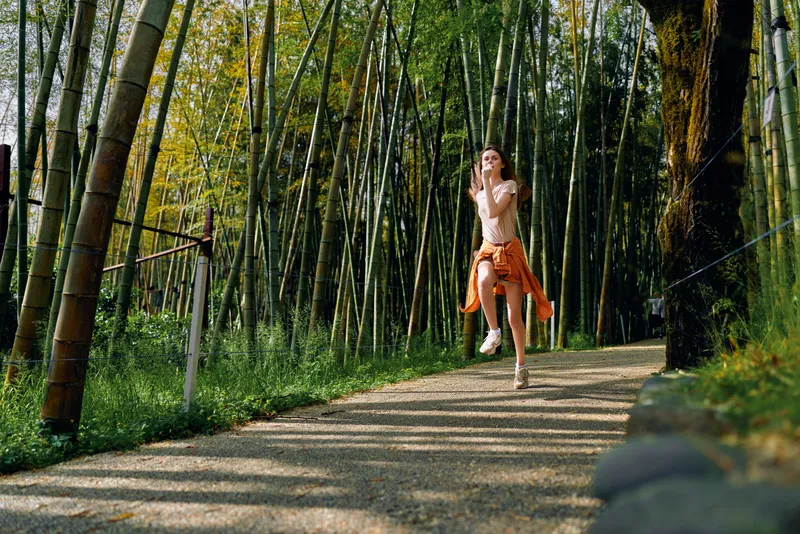 Woman running and jogging on a sunlit bamboo forest path, nature trail with gravel walkway, smiling and active in casual outfit with orange jacket tied, outdoor fitness and joyful movement.