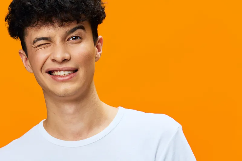 young man winking with friendly smile wearing white t-shirt against bright orange background, casual hairstyle and playful mood in studio lighting