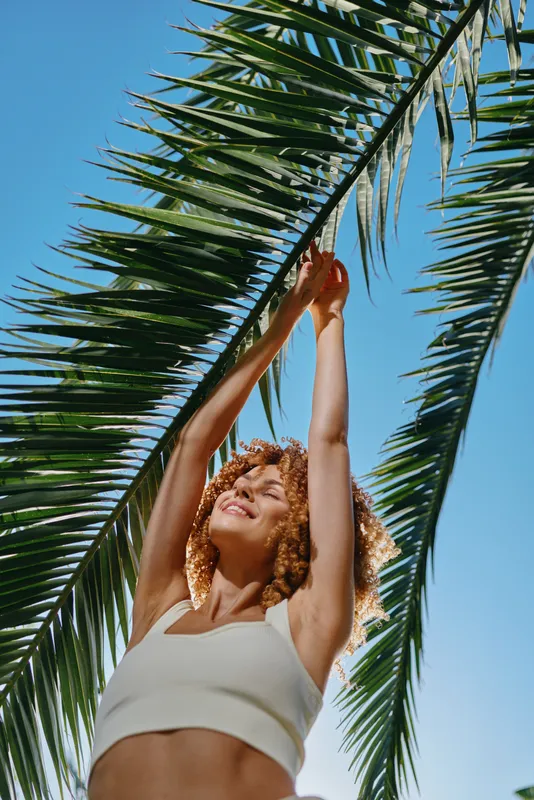 smiling woman with curly hair reaches up to green palm leaves under clear blue sky, wearing casual white top, sunlight highlighting joyful summer mood.