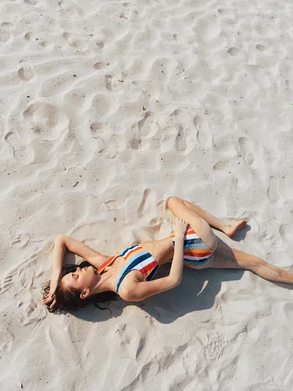 Female figure lying on white sandy beach wearing striped swimsuit, sunlit scene, relaxed pose, summer recreation, peaceful mood, warm tones and soft shadows.