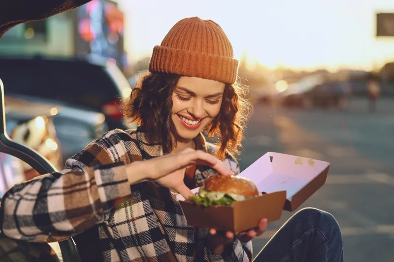Woman with beanie smiling while holding a burger in a takeout box by car at sunset, candid lifestyle portrait with authenticity, golden hour glow and emotional storytelling.