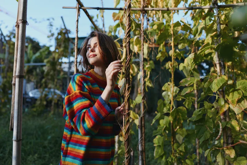 Woman in colorful striped sweater standing in garden among climbing vines, portrait of young person touching leaves, natural outdoors scene with warm sunlight and foliage.