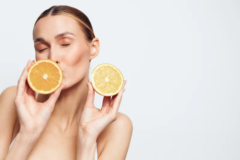 Young woman with clear skin holds two halves of citrus fruits close to her face, eyes closed, showing fresh natural beauty and healthy lifestyle on white background.